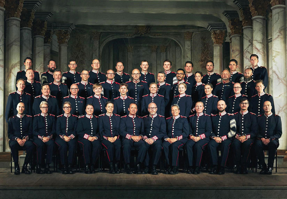 Musiker i uniform uppställda i fyra rader bakom varandra. Fotografi.