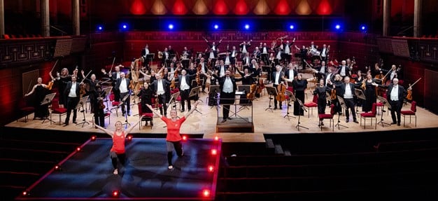 Två instruktörer som tränar framför en orkester. Fotografi.