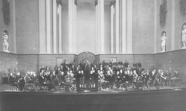 Scenen i Konserthusets stora sal sedd från parkettplats. Äldre gruppfotografi av dirigenten Václav Talich tillsammans med Kungliga Filharmonikerna, dåvarande Konsertföreningens orkester.